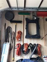 Photo of various extension cords in black and orange hanging on pegboard along with a broom, black lamp reflector, and other tools.