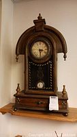 Front view of the wood framed mantle clock showing the curved top, clock face with Roman numerals, finials on top and sides, and lower drawer.