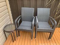 Two dark brown plastic rattan outdoor chairs next to one small round brown metal frame side table with glass top, placed on outdoor wooden flooring near wall and fence.