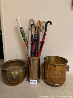 Photo showing two large brass pots and a brass umbrella stand filled with assorted umbrellas.