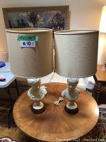 Two table lamps with off-white bust statue bases on wooden platforms, each topped with beige textured drum lampshades. Positioned on a round wooden table.