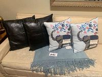Four pillows and a folded blue throw blanket displayed on couch. Two black leather pillows visible with stitched detail; two embroidered pillows have elephant, floral and butterfly motifs. Blue fringed throw blanket underneath all.