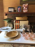 Full lot overview showing bun warmer, toaster, scale, recipe box, cookie press, cheese dip tray, pink glasses, and cake stand on table in front of fireplace.