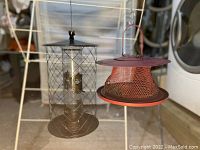 Photo showing two bird feeders hanging side by side: one cardinal red circular mesh feeder with a large capacity, the other a bronze metal cage style feeder for small birds.