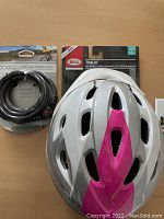 Front view of white and pink Bell Thalia helmet with LED cable lock coiled adjacent, both in packaging.