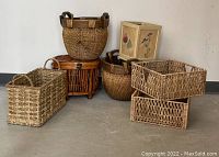 Seven assorted woven baskets in varying shapes and sizes placed on concrete floor, shown against white wall. Includes round, square, oval, rectangular shapes and a waste basket.