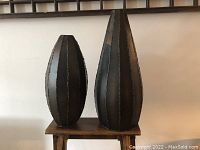 Photo showing two dark oblong metal vases with visible welded panel seams on a wooden stool against a white wall background.