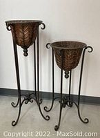 Photo shows two decorative metal plant holders on stands against a plain background. The taller holder is approximately 37 inches tall and 13 inches in diameter. The shorter holder is approximately 30 inches tall and 13 inches in diameter. Both have a leaf pattern and curled leg stands.