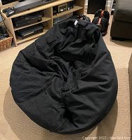 Black adult size bean bag chair with cotton cover placed on beige carpet in a living room setting, showing overall condition and shape.