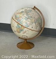Photo of an old fashioned world globe with a metal stand on concrete floor against a white wall.