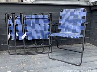 Four black metal framed lawn chairs with blue and white webbed seating and backrest. One chair is set up upright and the other three are stacked behind it against a dark wall background on wooden decking.