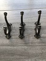 Photo of three large heavy metal coat hooks laid flat on a wood floor. Hooks have two curved prongs each and three mounting holes per piece. Dark finish with decorative mounting plate shape.