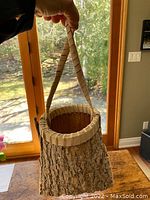 Photo shows a small Indigenous bark container with rough bark body, woven rim and handle held up by a hand in front of a window.