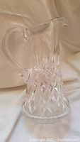 Photo showing a clear Waterford crystal pitcher with a diamond cut pattern and curved handle, set against a beige fabric background.