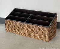 Wicker magazine rack with black plastic inner compartment dividers, photographed on a concrete floor against a neutral wall.