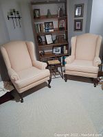 Photo showing two pale peach wingback chairs in a room with bookshelf and side table.
