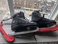 Side view of women's size 7 black figure skates placed on wooden surface by window with city view. Red blade guards attached to blades.