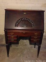 Front view of closed cherry wood secretary desk with carved shell motif on front panel and brass drawer pulls on lower drawers.
