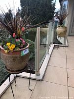 Two large ceramic planters filled with various flowering plants on black metal stands, placed on balcony with scenic background.