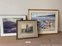 Three framed art pieces grouped together on floor showing colors and composition, includes architecture and landscape subjects.