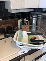 Wooden cutting boards at back with knife block holding knives, metal thermos, partly visible glass platter, and baking dishes in front with kitchen towels and utensils