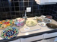 Wide view of 13 decorative plates and a lidded ceramic dish arranged on a counter surface.