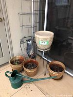 Overview photo showing several ceramic planter pots including large beige patterned pot on metal stand, green watering can, smaller brown pots, and gray plastic owl perched on a metal stand.