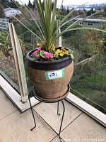 View of the large ceramic planter with dark rim on a black metal stand placed on a balcony with background of trees and buildings.