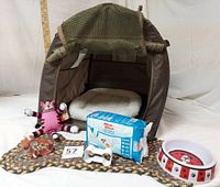 Overall lot showing khaki collapsible dog crate with attached white pillow, bone-shaped braided rug underneath, dog bowl, plush pink cat toy, cookie cutters, and disposable dog wraps.