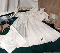 White christening gown with matching bonnet, stuffed unicorn security blanket, elephant motif tan and gray baby blanket, wooden animal wall art, and photo holder arranged on a dark green fabric background
