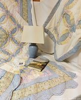 Photo showing quilt bedspread and sham draped over a surface, blue ceramic lamp with rectangular shade, and the Jane Austen book placed on the quilt.