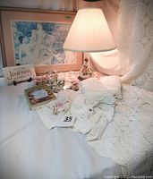 Full shot of crocheted coverlet draped with lamp, framed Renoir print, embroidered scarf and book visible.