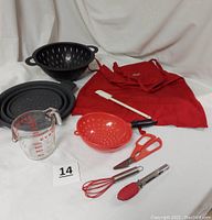 Photo showing all items in lot including Pampered Chef collapsible strainer, Pyrex measuring cup, red handheld strainer, black colander, mini whisk and tongs, scissors, silicone spatula, and red Wells Fargo apron.