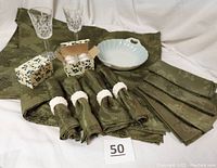 Photo showing green tablecloth with raised leaf design, eight matching napkins, eight white ceramic napkin rings in original boxes, vintage French-style china bowl, and two cut crystal stemmed wine glasses.