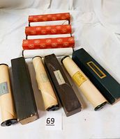 Six early 1900-1940s player piano rolls in original boxes, arranged on a white cloth background. Boxes have varying colors (black and orange) with visible branding and labels.