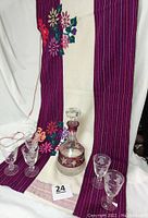 Photo showing the wine decanter and four of the five stemmed wine glasses arranged on a purple and cream embroidered table runner.