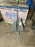 Full view of the metal boat anchor showing two large triangular flukes, central shank, and horizontal crossbar, placed on the floor with boxes behind it.