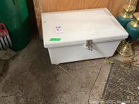Closed white marine grade fibreglass boat storage box with metal latch on front and visible scratches on exterior surface, placed on concrete floor near green trash container and brass lamps.
