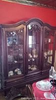 Front angled view of the wood and glass display cabinet showing three glass doors with visible contents inside, carved wood details on top and bottom, and one drawer at the base.