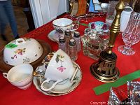 Lot contents including ceramic teacups, saucers, glass salt and pepper shakers, brass bell, and butter dish on wooden base with ceramic lid