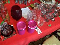 Overview of glasses, purple plastic cups with lids, vintage witch hazel bottle, large red glass vase, cloth napkins, and ashtrays on a red tablecloth