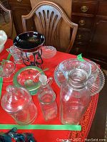 Glassware including ashtrays, sugar dish with spoon, pitcher with stopper (chip noted), jars and jugs arranged on a red cloth