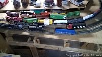 Overview of 25 HO scale model rail cars and locomotives arranged on wooden track with model buildings in background.
