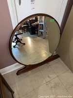 Front full view of large round mirror with wooden frame and wooden base stand, showing clear reflections and overall shape.