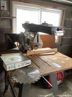 Radial arm saw mounted on wooden stand with circular saw blade. Boxed Dewalt 8 inch disc sander on table and additional saw blade beside it.