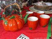 Photo showing the bright orange-red tomato shaped teapot with green leaf accents and woven bamboo handle together with four matching small cups on a red tablecloth.