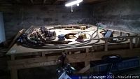 Rectangular plywood train platform with multiple loops of HO scale train track and some model buildings, elevated on wooden legs in a basement.
