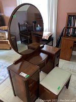 Wooden vanity with a large round mirror and fabric-covered stool beside it. Vanity has 4 drawers and a curved open space in the center.