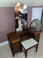 Full view of the wood vanity with mirror and fabric covered stool placed in front, showing overall condition and layout.