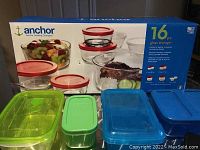 16 Piece Anchor Hocking glass storage containers set in original packaging with 4 used plastic containers in green and blue on a reflective table surface.